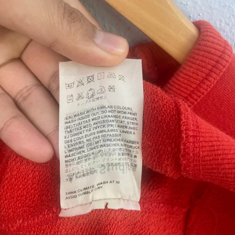 Op de afbeelding: Close-up van een rood kledingstuk met een wit etiket. Het etiket bevat wasvoorschriften in meerdere talen, met was- en droogsymbolen. De tekst bevat zinnen als "Wassen met dezelfde kleuren" en "Niet in de droger."