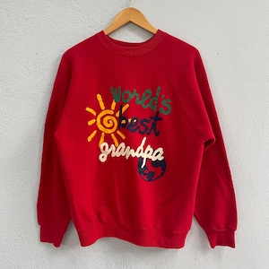 May include: A red sweatshirt featuring the text "World's Best Grandpa" in green, yellow, and white. The design includes a yellow sun and a blue and green globe. The sweatshirt is displayed on a wooden hanger.