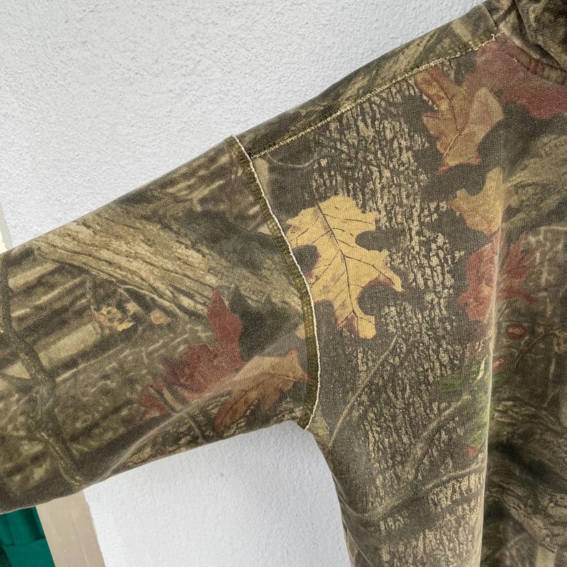 May include: Close-up of a camouflage-patterned long-sleeve shirt. The fabric features a mix of brown, green, and tan hues with leaf and branch details. The stitching is visible along the seams of the sleeve and shoulder.