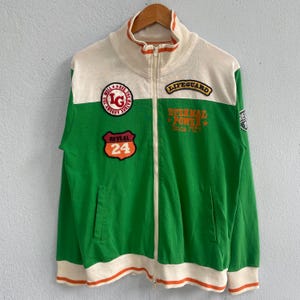 May include: Green and white zip-up jacket with orange accents. Features patches with text including "Lifeguard", "Eternal Power Since 1924", and "Reveal 24". The jacket has a high collar and two front pockets.
