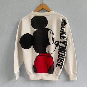 Puede incluir: Una sudadera de manga larga color crema con un gr&aacute;fico grande de Mickey Mouse en la parte posterior. El personaje aparece en negro, blanco y rojo, con el texto "MICKEY MOUSE" impreso verticalmente en letras negras negritas en el lado derecho.