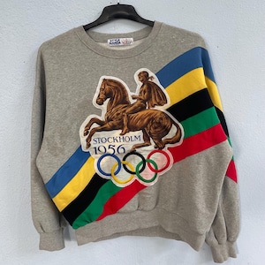 May include: A gray sweatshirt with a colorful diagonal stripe and a vintage Olympic design. The design features a rider on a horse, the text "STOCKHOLM 1956", and the Olympic rings. The sweatshirt has a crew neck and long sleeves.