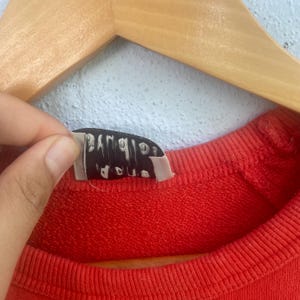 Op de afbeelding: Close-up van een rode sweatshirt met een geribbelde halslijn. Een zwart label met witte tekst is gedeeltelijk zichtbaar, vastgehouden door een hand. De sweatshirt hangt aan een houten hanger tegen een lichtblauwe muur.