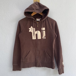 May include: A brown zip-up hoodie with a hood and front pockets. The word "hi" is printed on the front in a cream color, along with a flower and a heart-shaped design. The hoodie has a full zipper and a drawstring hood.