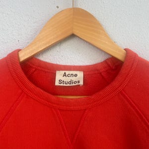 Op de afbeelding: Een rode crewneck sweatshirt met het label "Acne Studios". De sweatshirt hangt aan een houten hanger. De stof lijkt een zacht, gebreid materiaal te zijn.