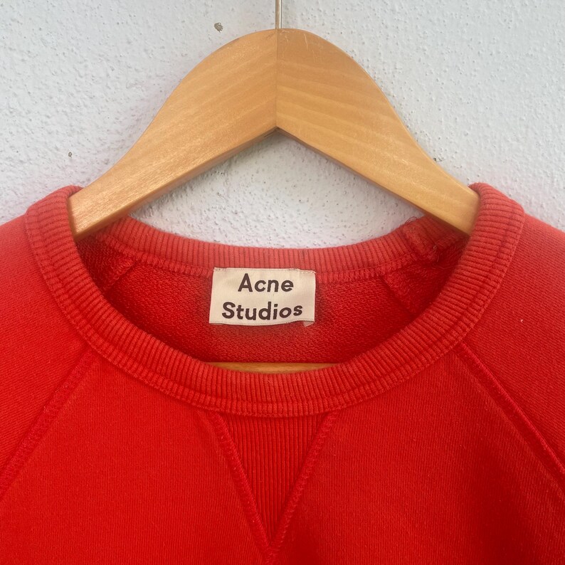 Op de afbeelding: Een rode crewneck sweatshirt met het label "Acne Studios". De sweatshirt hangt aan een houten hanger. De stof lijkt een zacht, gebreid materiaal te zijn.