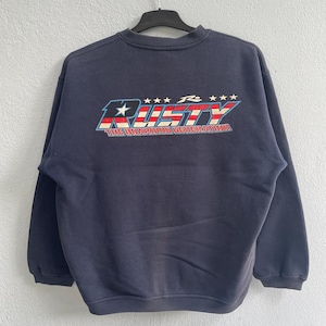 May include: Navy blue sweatshirt with the word "RUSTY" in a red, white, and blue design across the back. The text "THE BOARDING GENERATION" is below the word "RUSTY". The sweatshirt has a crew neck and long sleeves.