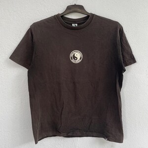 Puede incluir: Una camiseta marr&oacute;n oscuro con un dise&ntilde;o gr&aacute;fico redondo que presenta un s&iacute;mbolo del yin-yang y el texto "T.C. Surf Designs Stay Stoked". La camiseta est&aacute; colgada en una percha negra contra una pared gris claro.