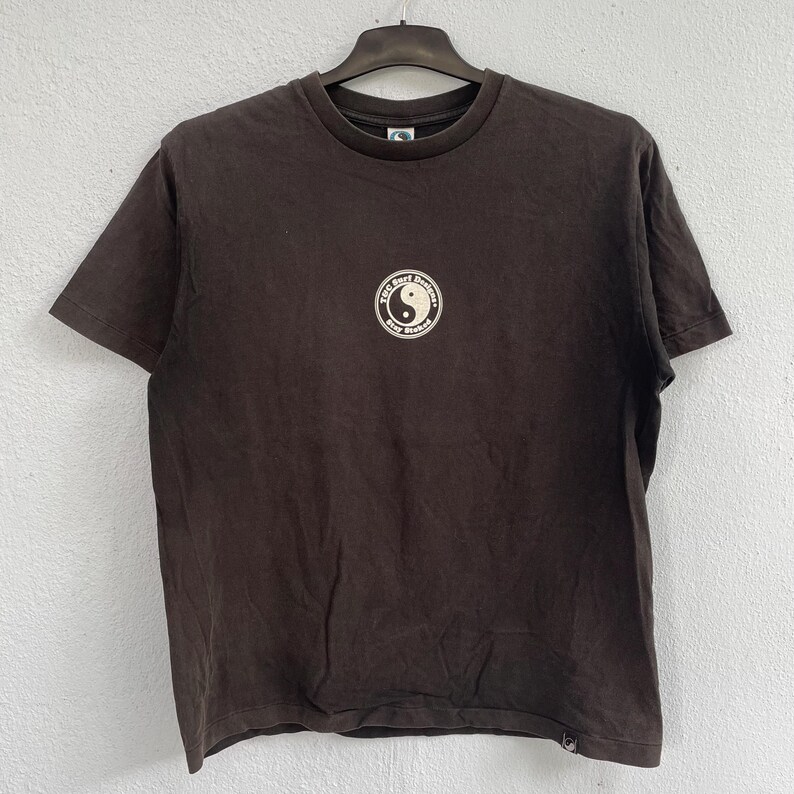 Puede incluir: Una camiseta marr&oacute;n oscuro con un dise&ntilde;o gr&aacute;fico redondo que presenta un s&iacute;mbolo del yin-yang y el texto "T.C. Surf Designs Stay Stoked". La camiseta est&aacute; colgada en una percha negra contra una pared gris claro.