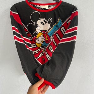 May include: Black long-sleeve shirt featuring Mickey Mouse holding a football. The shirt has red and white stripes on the sleeves and a red neckline and cuffs. The graphic includes the text "EFFORT" and "APTITUDE".