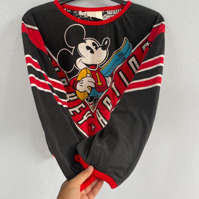 May include: Black long-sleeve shirt featuring Mickey Mouse holding a football. The shirt has red and white stripes on the sleeves and a red neckline and cuffs. The graphic includes the text "EFFORT" and "APTITUDE".