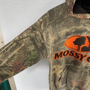 May include: A camouflage-patterned hoodie with the "Mossy Oak" logo in orange. The hoodie features a drawstring hood and a front pocket. The fabric has a mix of green, brown, and tan colors, creating a woodland camouflage design.
