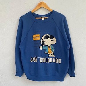 Peut inclure: Un sweat-shirt bleu &agrave; col rond arborant un motif du personnage de dessin anim&eacute; Snoopy portant des lunettes de soleil et une veste. Le motif inclut un panneau jaune indiquant Colorado Springs et le texte Joe Colorado imprim&eacute; en blanc en dessous.