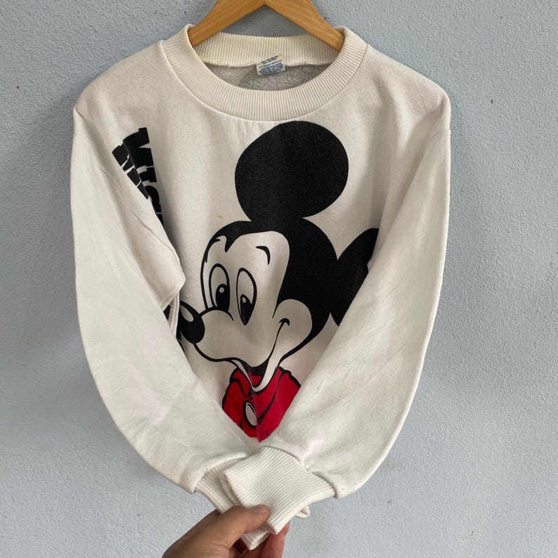 Puede incluir: Una sudadera blanca de manga larga con un gr&aacute;fico grande en blanco y negro de Mickey Mouse con una camisa roja. La sudadera tiene un cuello redondo acanalado y pu&ntilde;os acanalados. La palabra Mickey est&aacute; impresa en negro verticalmente en el lado derecho.