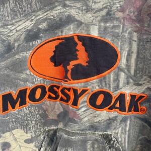May include: A camouflage-patterned hoodie with the "Mossy Oak" logo in orange and black. The logo features a tree silhouette within an oval. The hoodie has a drawstring hood and a front pocket.