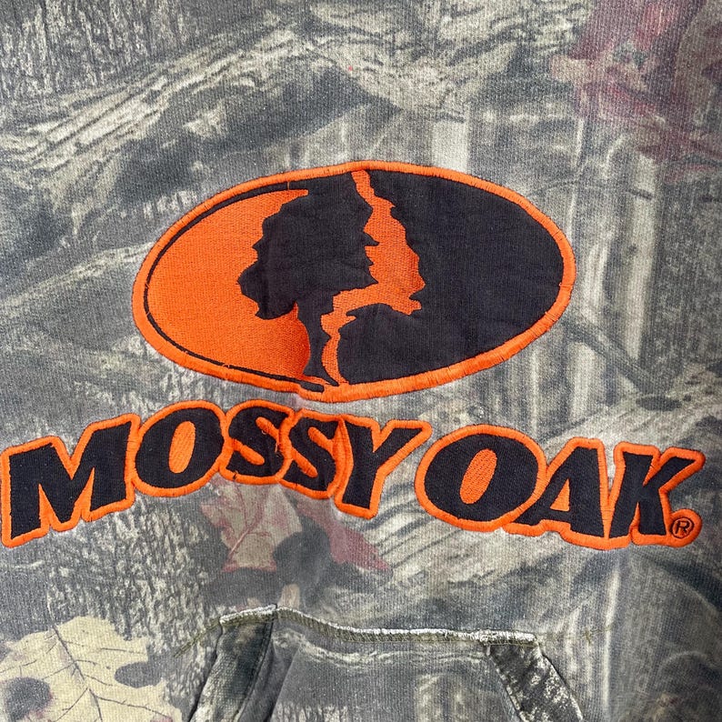 May include: A camouflage-patterned hoodie with the "Mossy Oak" logo in orange and black. The logo features a tree silhouette within an oval. The hoodie has a drawstring hood and a front pocket.