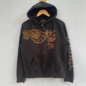 May include: Dark brown zip-up hoodie with a hood and front pockets. Features a gold dragon design on the front and sleeve, along with Japanese text. The hoodie has a drawstring and a wooden hanger.