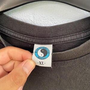 Puede incluir: Primer plano del cuello de una camiseta gris oscuro. La etiqueta presenta un s&iacute;mbolo de yin-yang dentro de un c&iacute;rculo azul, con el texto "T.U.C. Surf Designs Hawaii". La etiqueta tambi&eacute;n tiene la talla "-XL-".