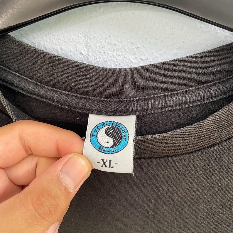 Puede incluir: Primer plano del cuello de una camiseta gris oscuro. La etiqueta presenta un s&iacute;mbolo de yin-yang dentro de un c&iacute;rculo azul, con el texto "T.U.C. Surf Designs Hawaii". La etiqueta tambi&eacute;n tiene la talla "-XL-".