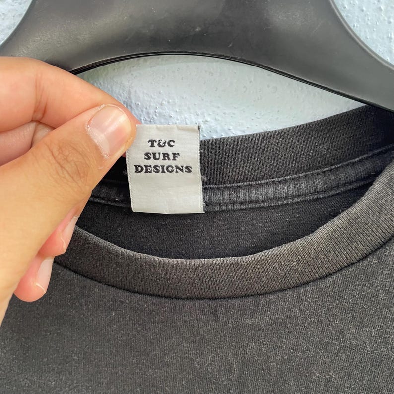 Puede incluir: Primer plano de un cuello de camiseta negra con una etiqueta blanca. La etiqueta dice "T&C SURF DESIGNS" en letras may&uacute;sculas negras. La camiseta est&aacute; hecha de una tela suave y oscura.