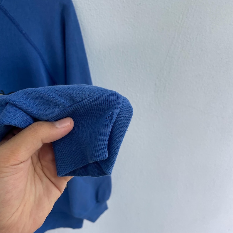Peut inclure: Un sweat-shirt bleu &agrave; manches longues dot&eacute; d'un poignet c&ocirc;tel&eacute;. Le tissu pr&eacute;sente un petit trou visible pr&egrave;s de la zone du poignet c&ocirc;tel&eacute;. Le v&ecirc;tement est con&ccedil;u avec des manches raglan et une couleur bleue unie sur toute la partie visible.