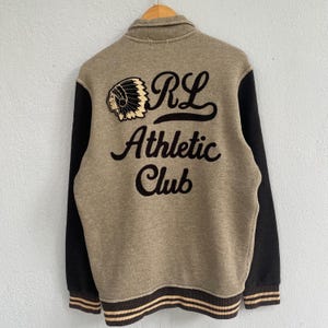 Op de afbeelding: Een beige en zwarte athletic club jas met de tekst "RL Athletic Club" geborduurd op de rug. De jas heeft zwarte mouwen, een knoopsluiting en een Native American hoofdlogo. De jas heeft een bruine en beige gestreepte bies.