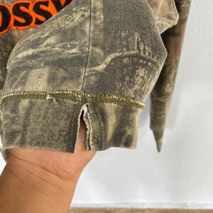May include: Close-up of a camouflage-patterned long-sleeved shirt. The cuff of the sleeve is visible, with a split at the side and a green stitched seam. The shirt has a faded, worn appearance. The word "OSS" is visible in orange.