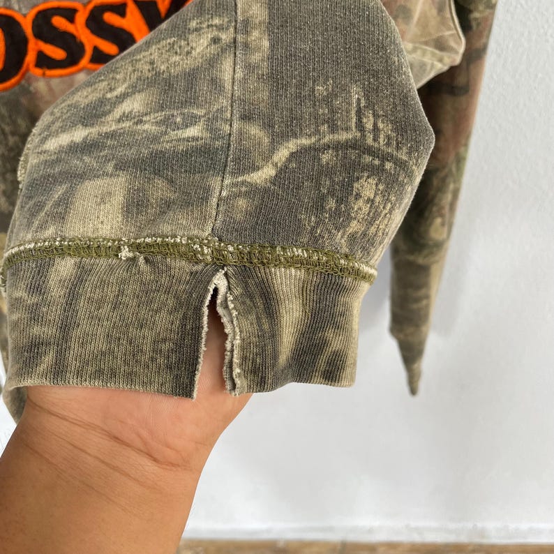 May include: Close-up of a camouflage-patterned long-sleeved shirt. The cuff of the sleeve is visible, with a split at the side and a green stitched seam. The shirt has a faded, worn appearance. The word "OSS" is visible in orange.