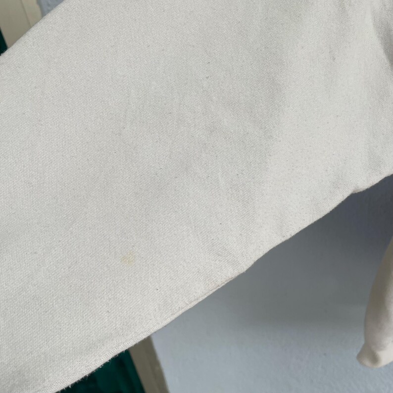 Puede incluir: Una vista de cerca de una manga de tela de color blanco roto con una textura tejida visible. El material parece ser un tejido resistente de color natural con una mancha amarillenta leve y tenue cerca del borde inferior de la tela.