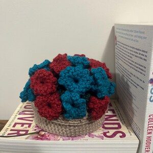 Puede incluir: Un arreglo de flores de ganchillo en una maceta marr&oacute;n. Las flores son rojas y azul verdoso.