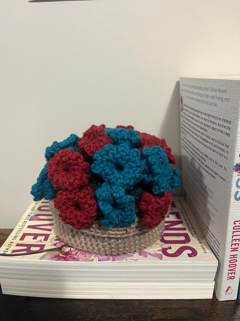 Puede incluir: Un arreglo de flores de ganchillo en una maceta marr&oacute;n. Las flores son rojas y azul verdoso.