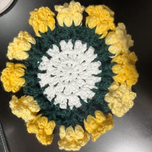 Puede incluir: Un posavasos de ganchillo con un centro blanco y un anillo exterior verde. El anillo exterior est&aacute; rodeado de flores de ganchillo amarillas.