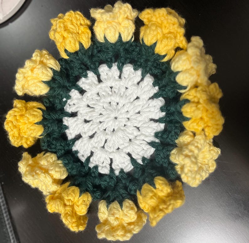 Puede incluir: Un posavasos de ganchillo con un centro blanco y un anillo exterior verde. El anillo exterior est&aacute; rodeado de flores de ganchillo amarillas.