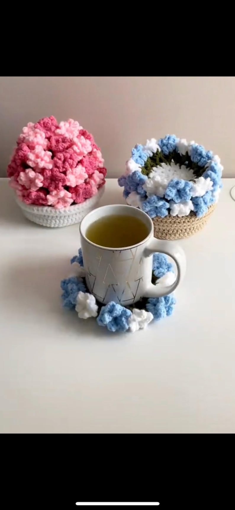 Puede incluir: Una taza de cer&aacute;mica blanca con un patr&oacute;n geom&eacute;trico dorado llena de t&eacute; verde se encuentra sobre un posavasos de ganchillo azul y blanco. Dos cestas de flores de ganchillo, una rosa y otra azul y blanca, est&aacute;n en el fondo.