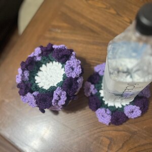 Puede incluir: Dos posavasos de ganchillo con un dise&ntilde;o floral morado y verde. Los posavasos est&aacute;n sobre una superficie de madera con una botella de agua transparente encima de uno de los posavasos.
