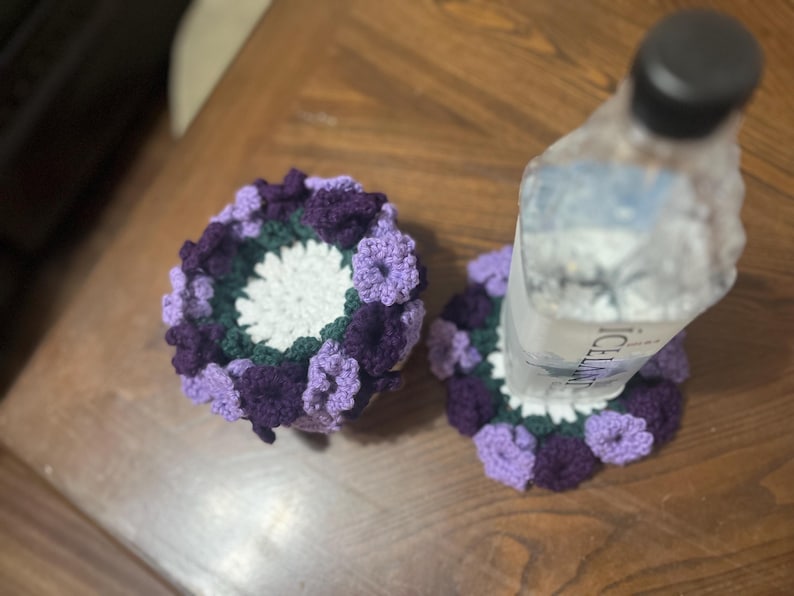 Puede incluir: Dos posavasos de ganchillo con un dise&ntilde;o floral morado y verde. Los posavasos est&aacute;n sobre una superficie de madera con una botella de agua transparente encima de uno de los posavasos.