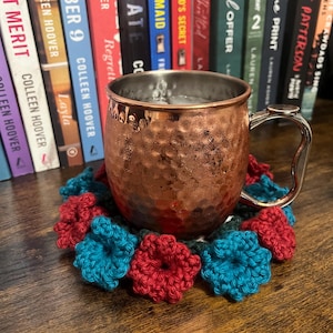 Puede incluir: Una taza de cobre con una bebida en su interior, sentada sobre un posavasos de ganchillo con flores rojas y azules. El posavasos est&aacute; sobre una mesa de madera. Hay libros en el fondo.