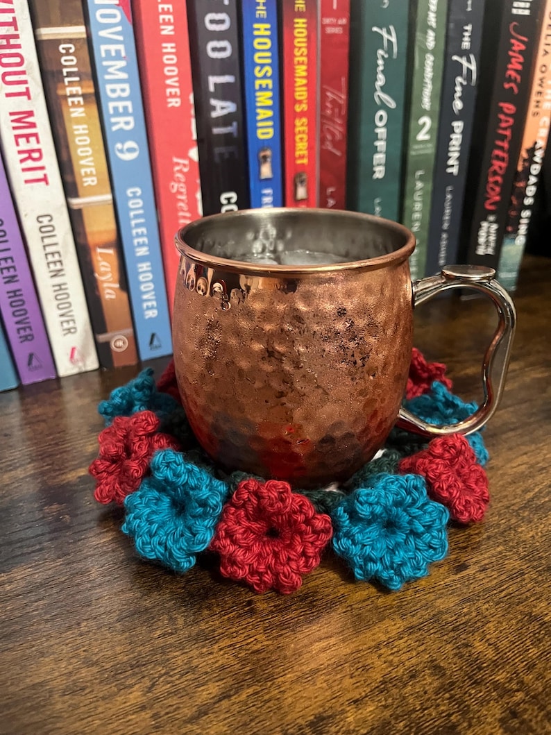 Puede incluir: Una taza de cobre con una bebida en su interior, sentada sobre un posavasos de ganchillo con flores rojas y azules. El posavasos est&aacute; sobre una mesa de madera. Hay libros en el fondo.