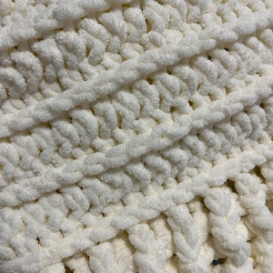 May include: A white chunky knit blanket with a textured, fluffy surface. The blanket is made with thick yarn and has a woven pattern.