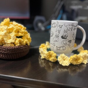 Puede incluir: Un posavasos de flores amarillas de ganchillo con una taza blanca con un dise&ntilde;o de gato blanco y negro. La taza est&aacute; sobre el posavasos.