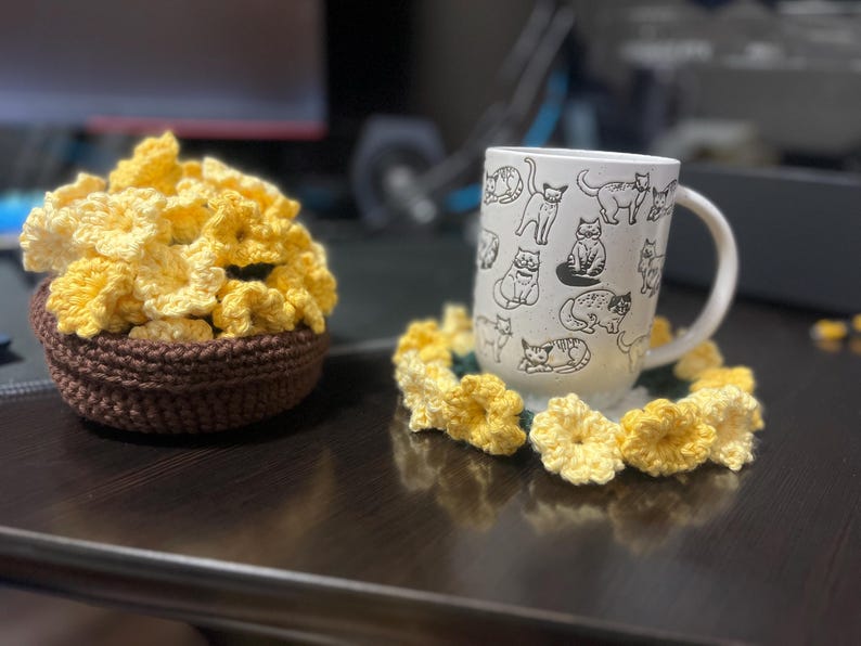 Puede incluir: Un posavasos de flores amarillas de ganchillo con una taza blanca con un dise&ntilde;o de gato blanco y negro. La taza est&aacute; sobre el posavasos.