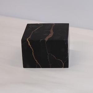 Könnte beinhalten: Ein schwarzer Marmorwürfel mit goldfarbenen Adern. Der Würfel ist eine solide, geometrische Form mit polierter Oberfläche. Die goldenen Adern bilden einen auffälligen Kontrast zum dunklen Hintergrund. Der Würfel ist ein dekoratives Objekt.