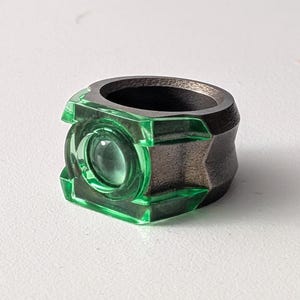 May include: A ring featuring a dark metallic band and a translucent green rectangular top section. The center of the green component contains a circular, raised, clear glass-like element, creating a distinct layered appearance.