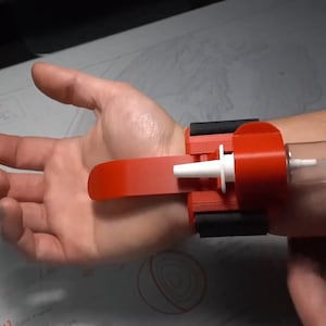 Può includere: Un dispositivo rosso e nero da polso con un componente cilindrico trasparente. Il dispositivo ha una leva rossa e un ugello bianco. Il dispositivo è tenuto in posizione con cinghie nere.