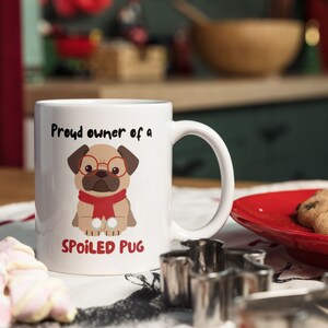 K&ouml;nnte beinhalten: Wei&szlig;e Keramik-Tasse mit Henkel, mit einem Cartoon-Mops mit Brille und rotem Schal. Auf der Tasse steht der Text "Proud owner of a SPOILED PUG". Ein roter Teller mit Keksen befindet sich neben der Tasse.