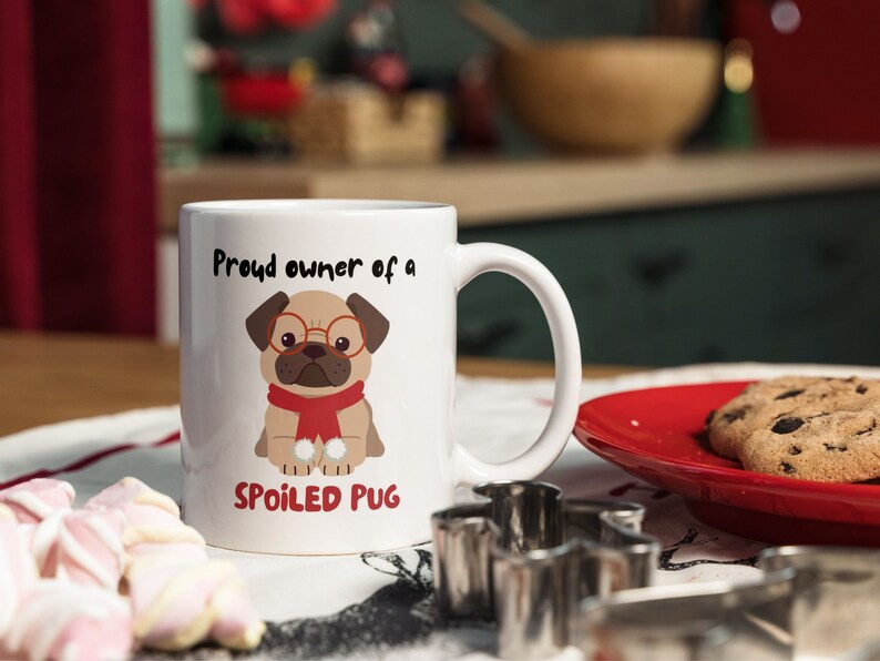 K&ouml;nnte beinhalten: Wei&szlig;e Keramik-Tasse mit Henkel, mit einem Cartoon-Mops mit Brille und rotem Schal. Auf der Tasse steht der Text "Proud owner of a SPOILED PUG". Ein roter Teller mit Keksen befindet sich neben der Tasse.