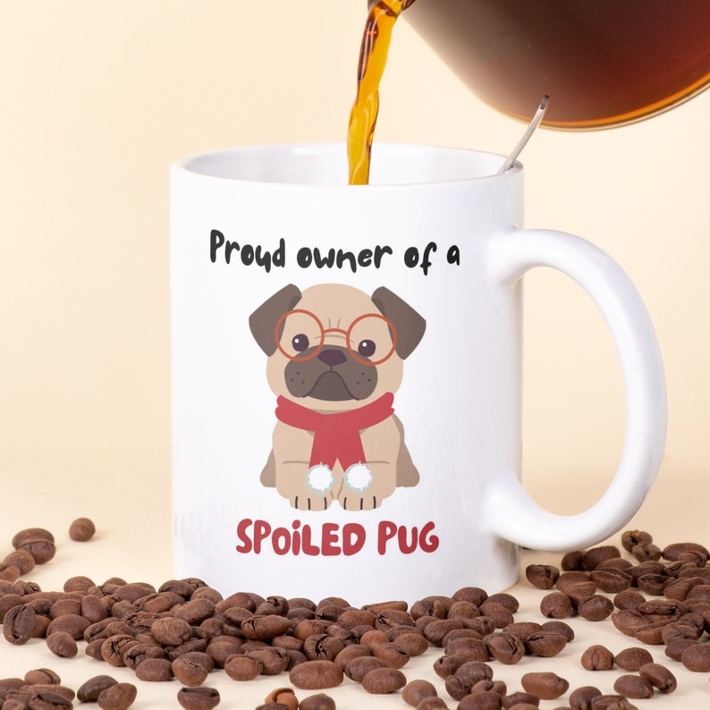 K&ouml;nnte beinhalten: Wei&szlig;e Keramik-Tasse mit einem Mops-Motiv, das eine Brille und einen roten Schal tr&auml;gt. Auf der Tasse steht "Proud owner of a SPOILED PUG". Kaffee wird in die Tasse gegossen, umgeben von Kaffeebohnen.