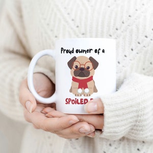 K&ouml;nnte beinhalten: Wei&szlig;e Keramiktasse mit einem Cartoon-Mops, der eine Brille und einen roten Schal tr&auml;gt. Auf der Tasse steht in schwarzer Schrift "Proud owner of a SPOILED pug". Die Tasse wird von einer Person gehalten, die einen cremefarbenen Pullover tr&auml;gt.