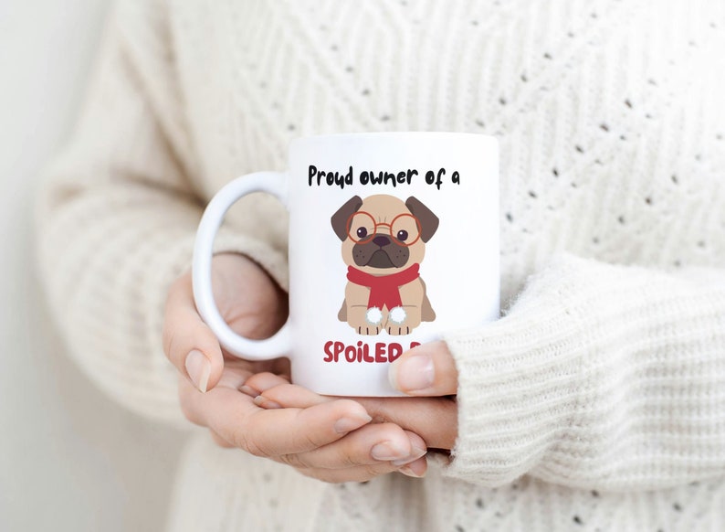 K&ouml;nnte beinhalten: Wei&szlig;e Keramiktasse mit einem Cartoon-Mops, der eine Brille und einen roten Schal tr&auml;gt. Auf der Tasse steht in schwarzer Schrift "Proud owner of a SPOILED pug". Die Tasse wird von einer Person gehalten, die einen cremefarbenen Pullover tr&auml;gt.