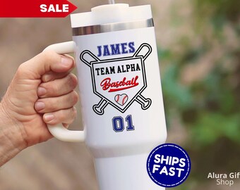 May include: White insulated tumbler with a handle, featuring a baseball-themed design. The design includes the name "JAMES" in blue, "TEAM ALPHA Baseball" in a shield, a baseball graphic, and the number "01". A "SALE" tag is visible.